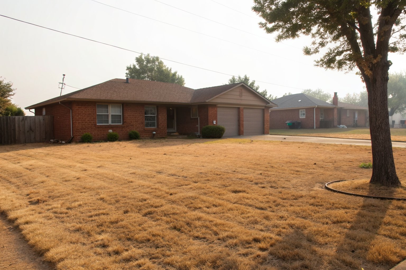 Brown, sun-dried patchy lawn in front of a one-story brick ranch house under a hazy summer sky